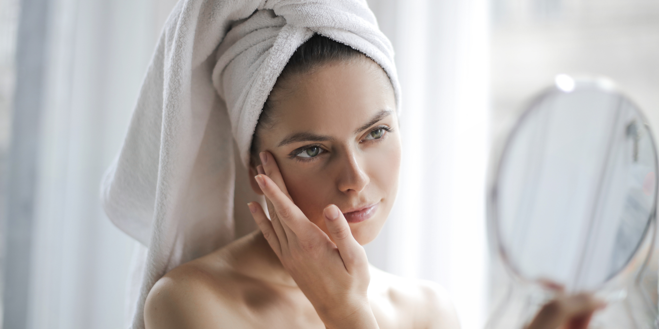 woman with her hair up in a towel, touching her face and looking in mirror
